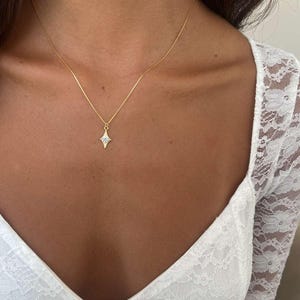 May include: A delicate gold necklace featuring a small, diamond-shaped pendant with a central gemstone. The pendant hangs from a thin gold chain. The necklace is shown against a skin tone background.