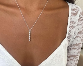 May include: A delicate silver necklace featuring a vertical row of four clear, sparkling gemstones. The necklace hangs from a fine chain, creating a minimalist and elegant design. The gemstones are evenly spaced and catch the light, adding a touch of glamour.