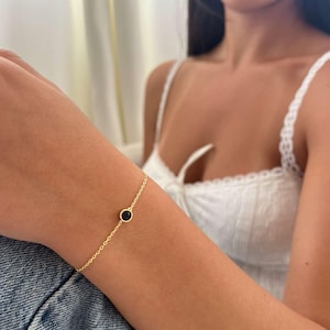 Op de afbeelding: Een fijne gouden armband met een kleine, ronde zwarte steen. De armband heeft een eenvoudig kettingontwerp en weerspiegelt een minimalistische esthetiek. Het sieraad wordt om een pols gedragen, tegen een achtergrond van denim en een witte top.