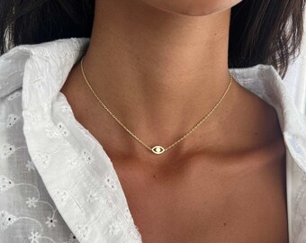 May include: A gold chain necklace with a small evil eye pendant. The necklace is worn by a person with dark hair and a white shirt.