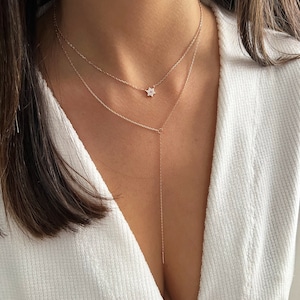 K&ouml;nnte beinhalten: Eine mehrreihige goldene Halskette mit einem kleinen Sternanh&auml;nger. Die Kette wird von einer Person mit langen braunen Haaren und einem wei&szlig;en Hemd getragen.