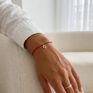 Op de afbeelding: Een dunne rode koordarmband met een kleine, zilverkleurige Davidster-bedel ingelegd met kleine doorzichtige steentjes, gecombineerd met een eenvoudige, gladde zilverkleurige ring om de vinger.
