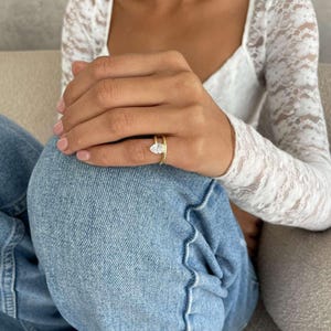 May include: A gold ring featuring a pear-cut diamond. The ring sits on a thin gold band. The person is wearing a white lace top and blue jeans.