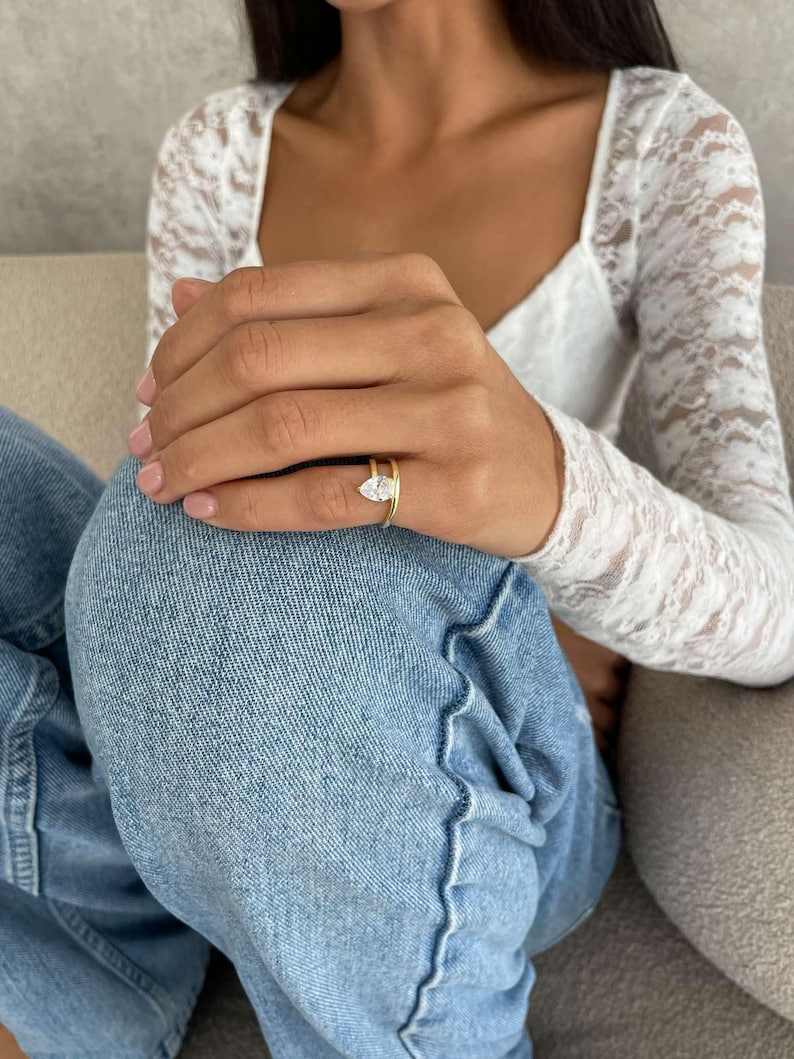 May include: A gold ring featuring a pear-cut diamond. The ring sits on a thin gold band. The person is wearing a white lace top and blue jeans.