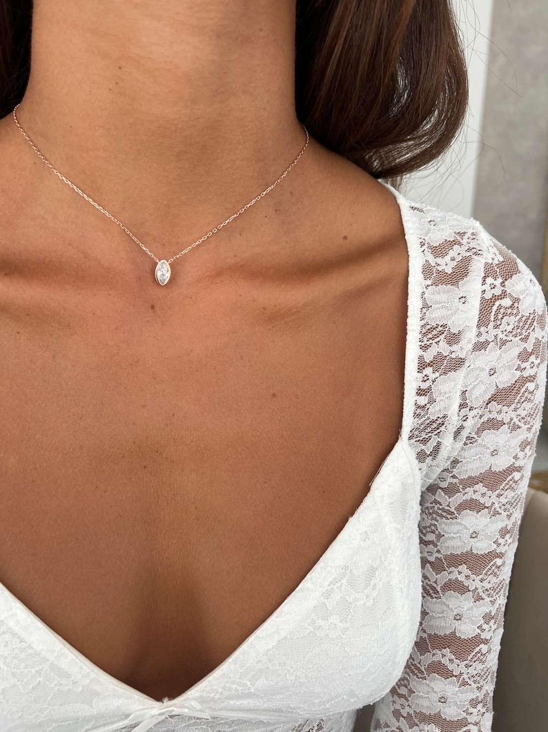 May include: A delicate rose gold necklace featuring a marquise-cut diamond pendant. The pendant is suspended from a fine chain, creating a minimalist and elegant look. The necklace is shown against a backdrop of a white lace top.