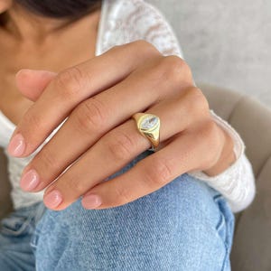 May include: A gold signet ring featuring a clear, marquise-cut gemstone. The ring has a polished finish and is worn on a finger. The background includes a white top and blue denim jeans.
