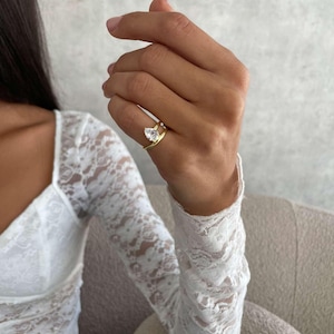 May include: A gold ring featuring a clear, pear-cut gemstone. The ring is worn on a hand, with a white lace sleeve in the background. The ring's band has a distinctive split design.