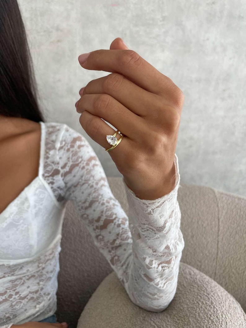 May include: A gold ring featuring a clear, pear-cut gemstone. The ring is worn on a hand, with a white lace sleeve in the background. The ring's band has a distinctive split design.