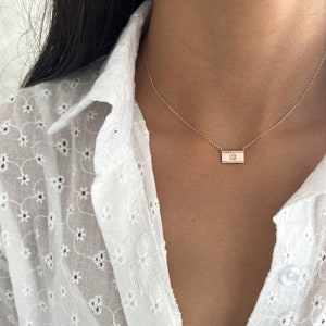 May include: A delicate gold necklace with a small rectangular pendant featuring a leaf design. The necklace is worn by a person with long black hair and a white blouse.