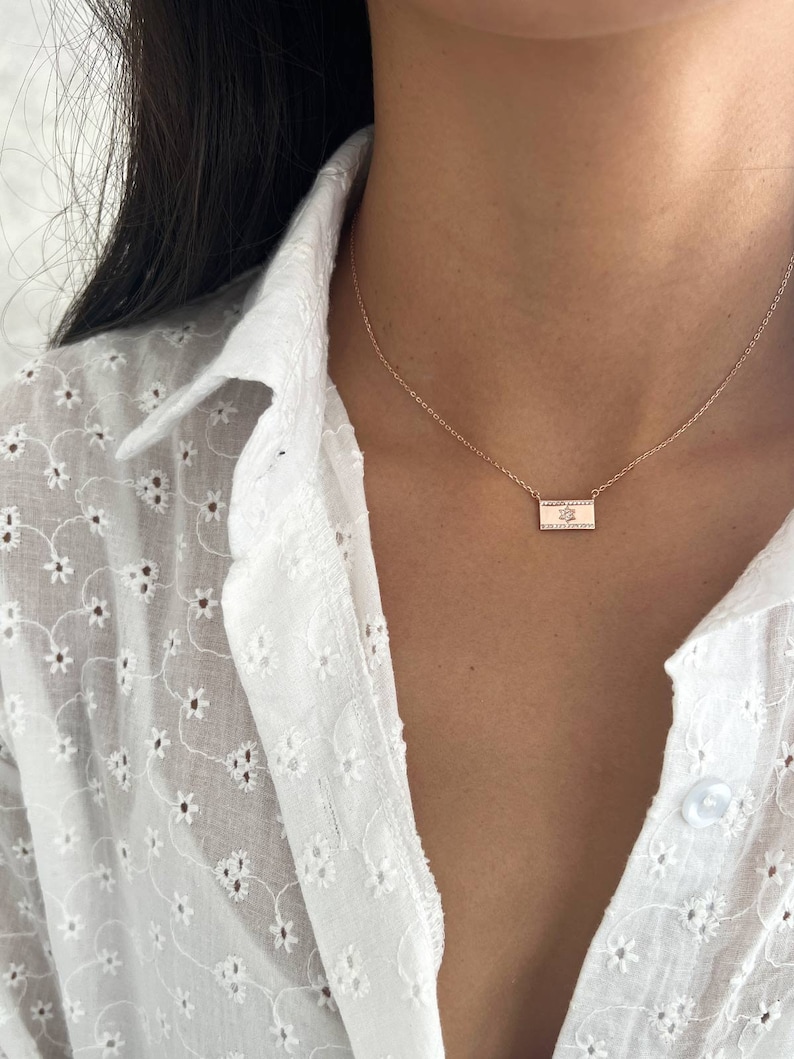 May include: A delicate gold necklace with a small rectangular pendant featuring a leaf design. The necklace is worn by a person with long black hair and a white blouse.