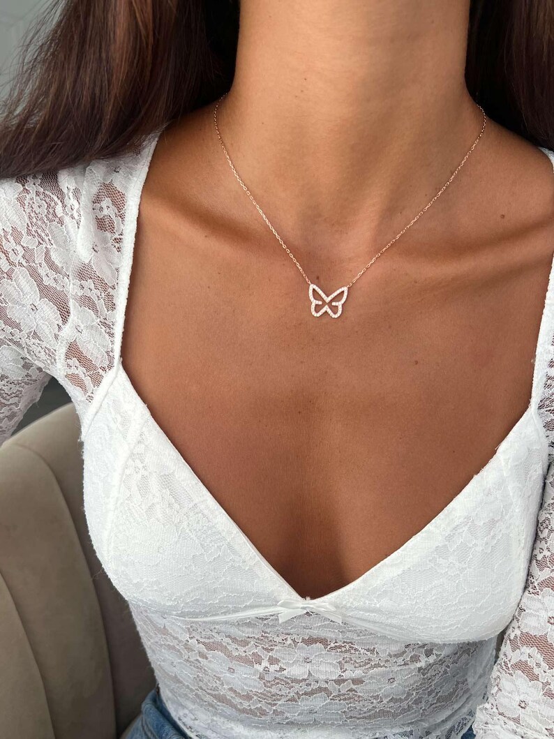 May include: A delicate gold-colored butterfly pendant necklace. The butterfly design is open and airy, suspended from a fine chain. The necklace is worn against a white lace top.