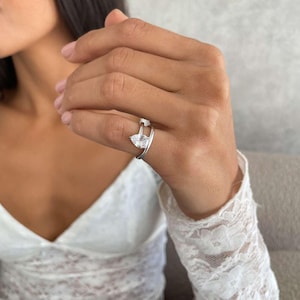 May include: A silver ring featuring a pear-cut, clear gemstone. The ring has a split shank design. The ring is on a person's finger, with a white lace sleeve in the background. The ring is a piece of jewellery.