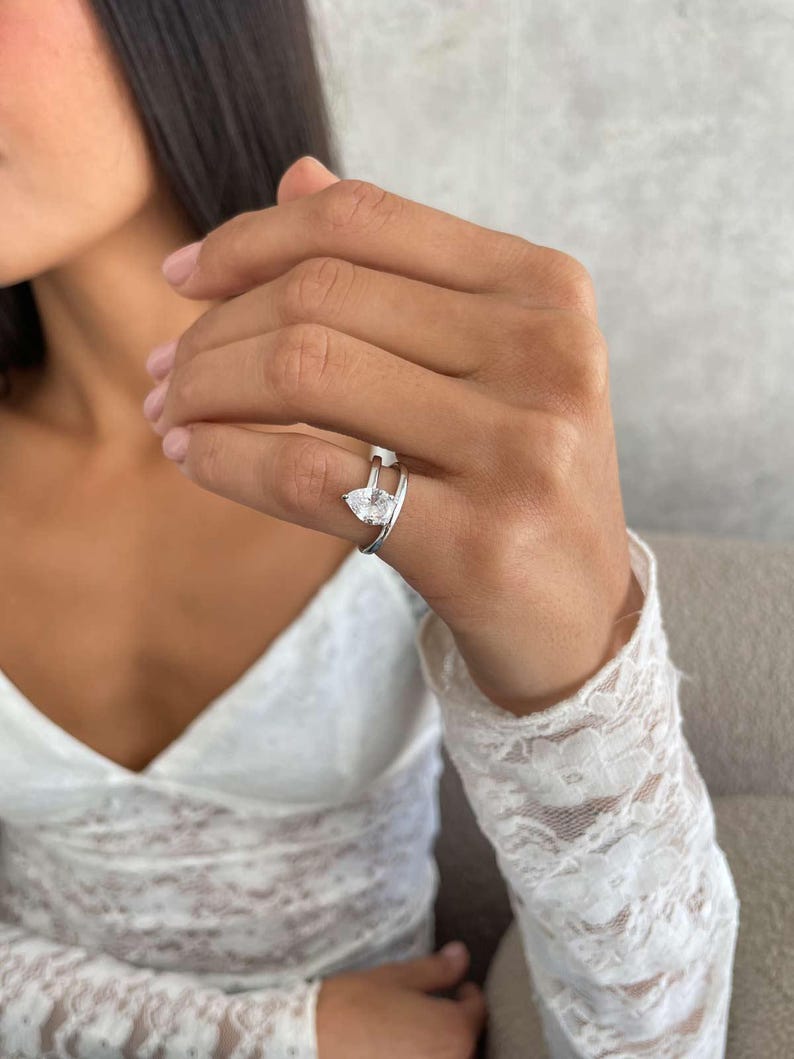 May include: A silver ring featuring a pear-cut, clear gemstone. The ring has a split shank design. The ring is on a person's finger, with a white lace sleeve in the background. The ring is a piece of jewellery.