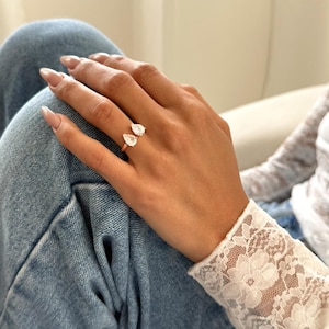 Puede incluir: Un anillo de oro rosa con dos gemas claras en forma de pera. El anillo se lleva en una mano con uñas largas y cuidadas. El fondo incluye unos vaqueros azules y una manga de encaje blanco.