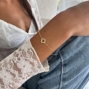 Op de afbeelding: Een delicate gouden armband met een klavertjevier bedel met heldere stenen. De armband wordt om een arm gedragen, met een witte kanten mouw en een blauwe spijkerbroek.