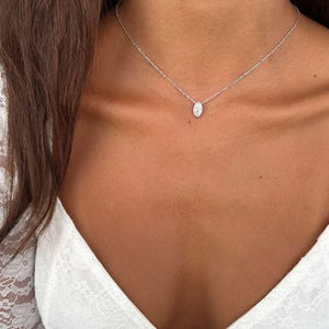 May include: A delicate silver necklace with a small, oval-shaped pendant. The pendant is set with a clear, sparkling stone. The necklace is worn on a person's neck, against a white lace top.