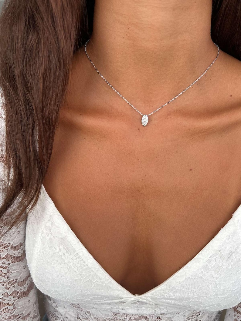 May include: A delicate silver necklace with a small, oval-shaped pendant. The pendant is set with a clear, sparkling stone. The necklace is worn on a person's neck, against a white lace top.