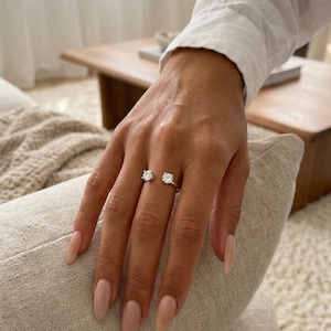 Op de afbeelding: Een zilveren ring met twee heldere, ronde edelstenen. De ring wordt gedragen aan een hand met lange, gepolijste nagels. De hand rust op een beige kussen, met een witte mouw op de achtergrond.