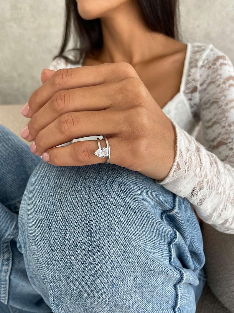 May include: A silver ring featuring a pear-cut diamond and a split shank. The ring is worn on a finger, highlighting its elegant and contemporary design. The diamond glitters, reflecting light.