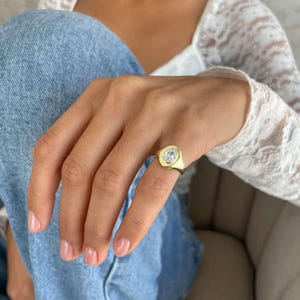 Può includere: Un anello con sigillo dorato con una pietra trasparente di forma ovale. L'anello è indossato su un dito, con uno sfondo in denim blu chiaro. L'incastonatura dell'anello è di colore oro lucido.