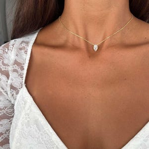 May include: A delicate gold necklace featuring a marquise-cut diamond pendant. The pendant hangs from a fine chain, resting gracefully on the neckline. The model is wearing a white lace top.