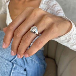 May include: A polished silver ring featuring a clear, oval gemstone. The ring is set in a smooth silver band. The design is simple and elegant, suitable for everyday wear or special occasions. The ring is worn on a finger.