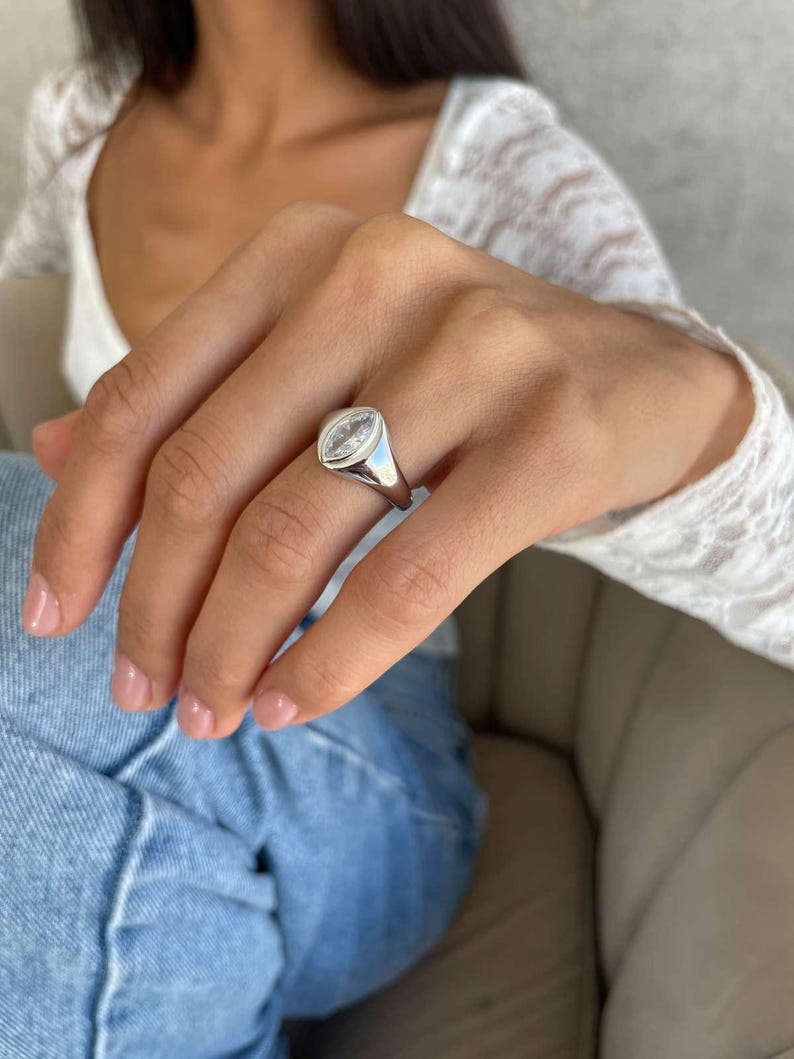 May include: A polished silver ring featuring a clear, oval gemstone. The ring is set in a smooth silver band. The design is simple and elegant, suitable for everyday wear or special occasions. The ring is worn on a finger.