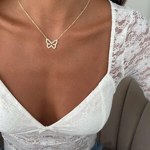 May include: A delicate gold-colored butterfly pendant necklace. The butterfly design is open and airy, suspended from a fine chain. The necklace is worn against a backdrop of a white lace top and blue jeans.