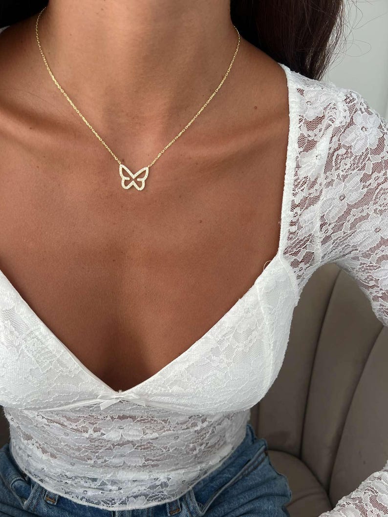 May include: A delicate gold-colored butterfly pendant necklace. The butterfly design is open and airy, suspended from a fine chain. The necklace is worn against a backdrop of a white lace top and blue jeans.