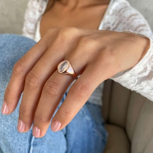 May include: Rose gold ring featuring a clear, oval-cut stone. The ring has a polished band and a bezel setting. A classic piece of jewellery, suitable for everyday wear or special occasions.