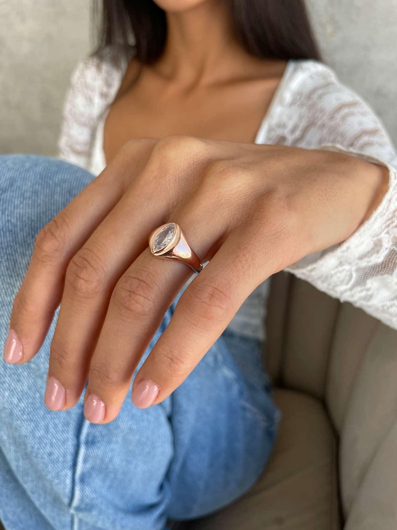 May include: Rose gold ring featuring a clear, oval-cut stone. The ring has a polished band and a bezel setting. A classic piece of jewellery, suitable for everyday wear or special occasions.