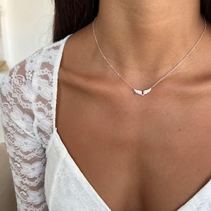 May include: A delicate necklace with a small wing pendant. The necklace is gold-toned, and the wings are adorned with small, sparkling stones. The necklace is worn on a person's neck, against a white lace top.