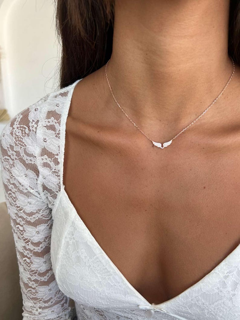 May include: A delicate necklace with a small wing pendant. The necklace is gold-toned, and the wings are adorned with small, sparkling stones. The necklace is worn on a person's neck, against a white lace top.