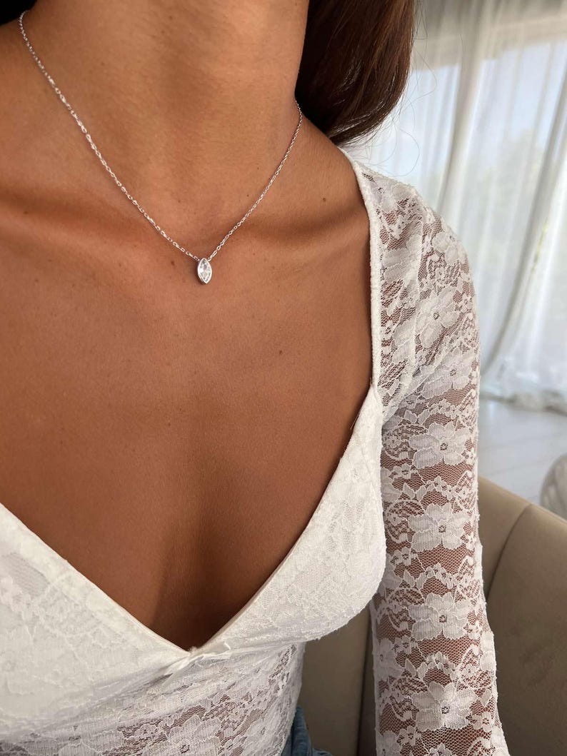 May include: A delicate silver necklace featuring a marquise-cut pendant. The pendant is suspended from a fine chain. The necklace is shown against a white lace top.
