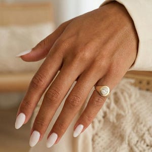 Op de afbeelding: Een gouden zegelring met een grote, ovaalvormige, heldere edelsteen wordt gedragen aan een hand met witte nagellak. De ring is het middelpunt, tegen een neutrale achtergrond van crèmekleurige kleding en een gebreide textuur.