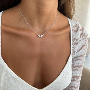 May include: A delicate silver necklace featuring a wing-shaped pendant. The necklace is worn on a person's neck, with the pendant resting near the collarbone. The person is wearing a white lace top.