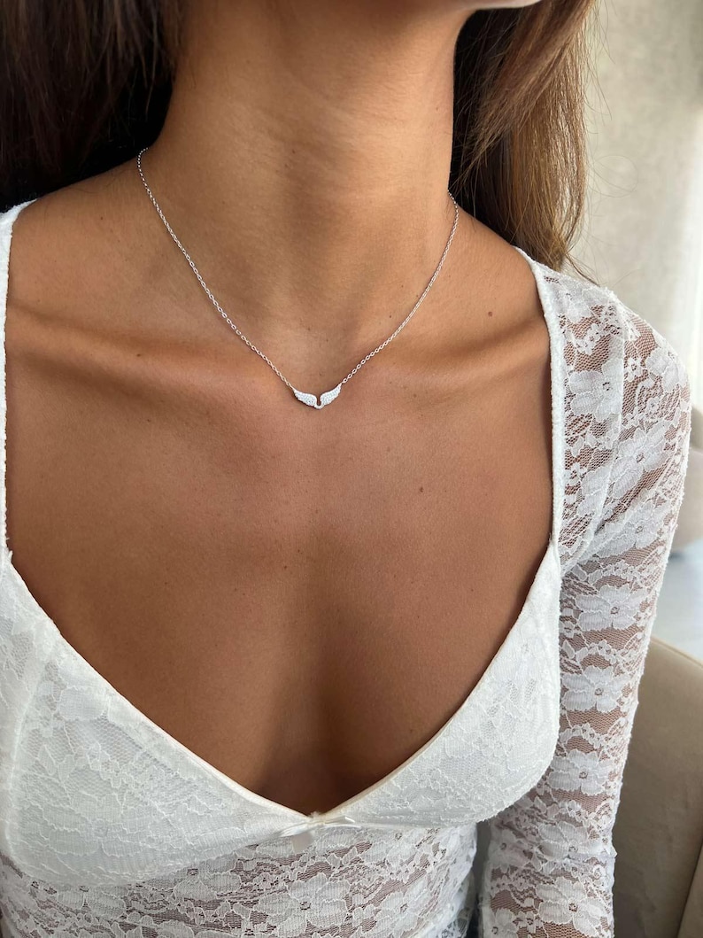 May include: A delicate silver necklace featuring a wing-shaped pendant. The necklace is worn on a person's neck, with the pendant resting near the collarbone. The person is wearing a white lace top.
