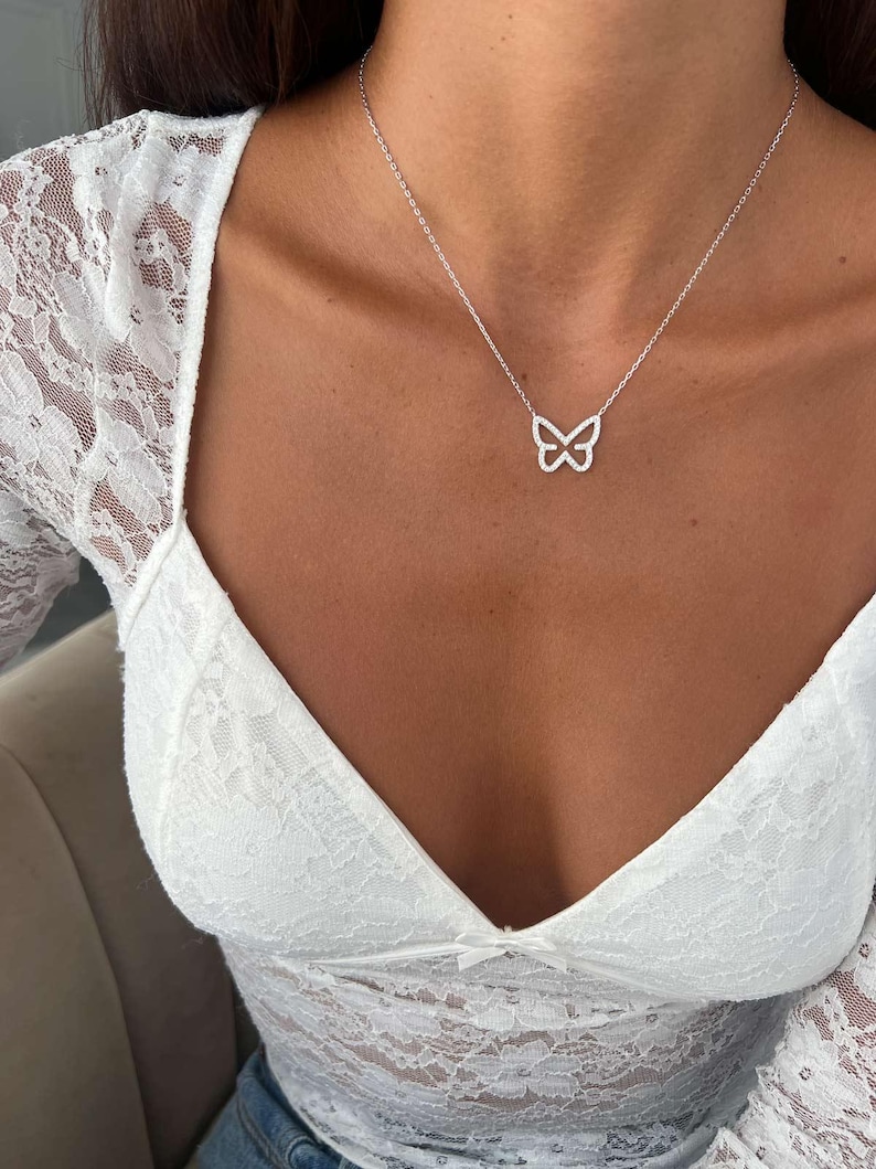 May include: A silver butterfly pendant necklace. The butterfly design is encrusted with small, sparkling stones. The necklace is worn on a person with a white lace top.