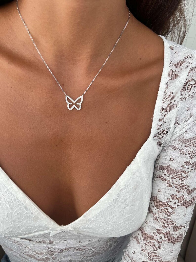 May include: A silver butterfly pendant necklace. The pendant is a delicate openwork design in the shape of a butterfly. The necklace is worn on a person with a white lace top.