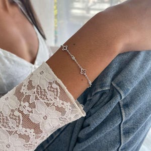 May include: A delicate silver bracelet featuring four-leaf clover charms and small round accents. The bracelet is worn on an arm, paired with a white lace sleeve and blue denim jeans. The jewelry is a subtle yet elegant accessory.