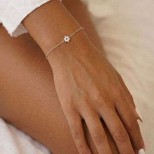 May include: A delicate silver bracelet featuring a star-shaped charm with a subtle, sparkling design. The bracelet is worn on a wrist, showcasing its fine chain and the intricate details of the star. The overall aesthetic is minimalist and elegant.