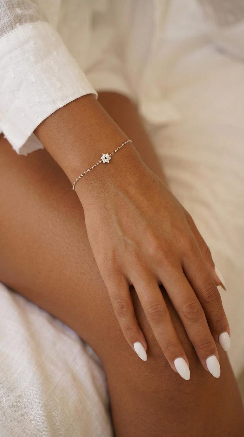 May include: A delicate silver bracelet featuring a star-shaped charm with a subtle, sparkling design. The bracelet is worn on a wrist, showcasing its fine chain and the intricate details of the star. The overall aesthetic is minimalist and elegant.