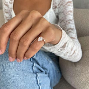 May include: A rose gold ring featuring a clear, pear-cut gemstone. The ring is worn on a finger, with a white lace top and blue jeans visible. The band is slender, and the gemstone is held in place by prongs.