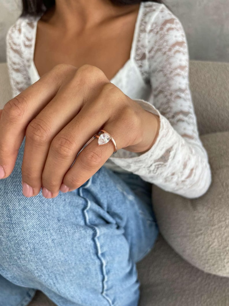 May include: A rose gold ring featuring a clear, pear-cut gemstone. The ring is worn on a finger, with a white lace top and blue jeans visible. The band is slender, and the gemstone is held in place by prongs.