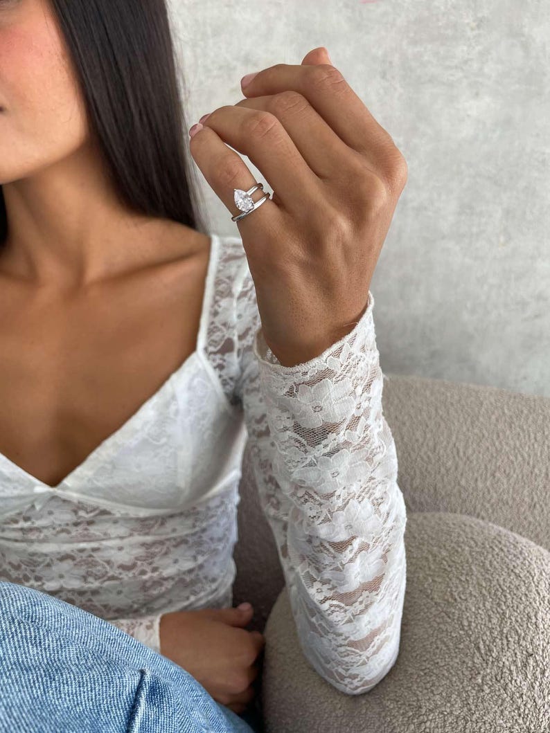May include: A silver ring with a pear-shaped gemstone is worn on a hand. The ring has a double band design. The person is wearing a white lace top and blue jeans. The background is a neutral colour.