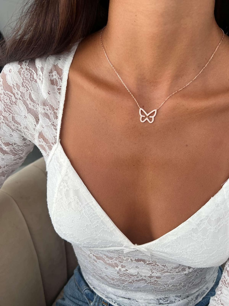 May include: A delicate rose gold butterfly pendant necklace. The butterfly design is encrusted with small, sparkling stones. The necklace is worn on a person wearing a white lace top.