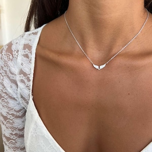 May include: A silver necklace with a wing pendant. The necklace is delicate and hangs on a person's neck. The pendant is made of small, sparkling stones. The person is wearing a white lace top.