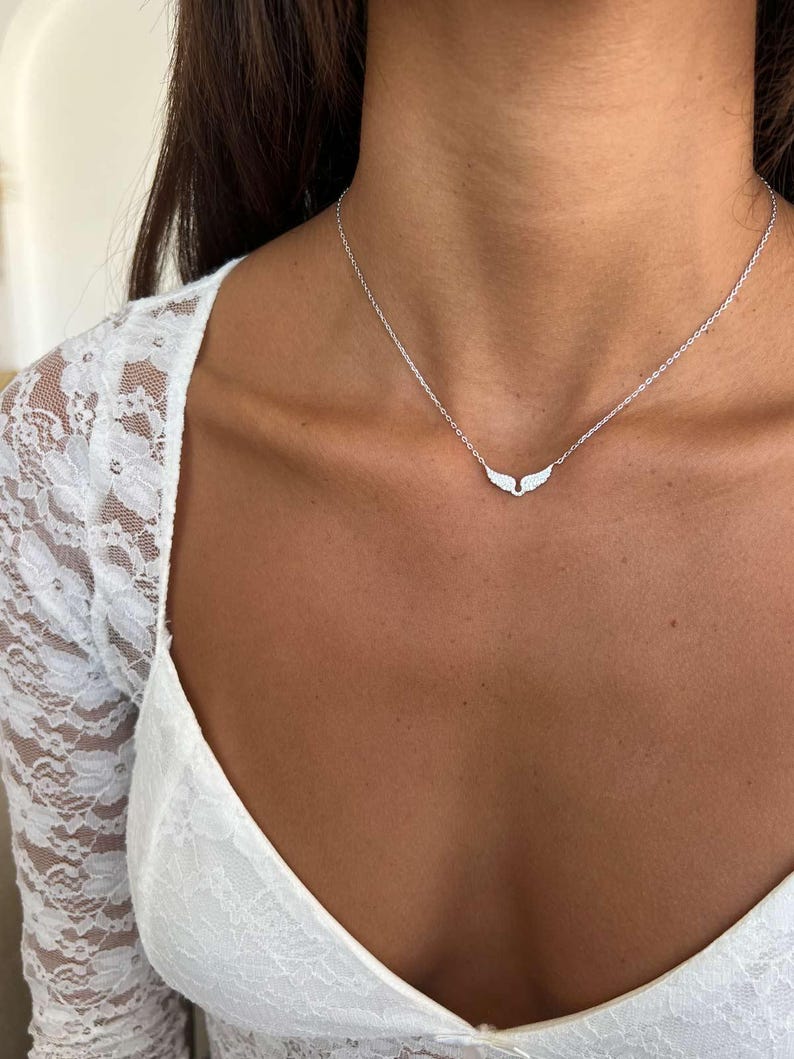 May include: A silver necklace with a wing pendant. The necklace is delicate and hangs on a person's neck. The pendant is made of small, sparkling stones. The person is wearing a white lace top.