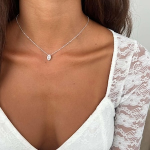 May include: A delicate silver necklace featuring a small, oval pendant. The pendant is centered on a fine chain, creating a minimalist and elegant design. The necklace is worn against a backdrop of a white lace top.