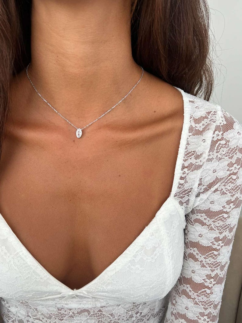 May include: A delicate silver necklace featuring a small, oval pendant. The pendant is centered on a fine chain, creating a minimalist and elegant design. The necklace is worn against a backdrop of a white lace top.
